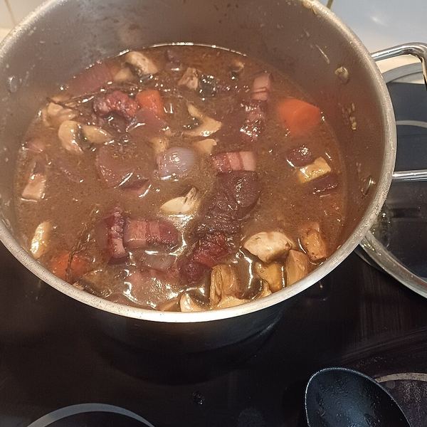 A pot of beef stew part-way through cooking, the liquid thin and clear rather than thickened, with beef, bacon and mushrooms in it.