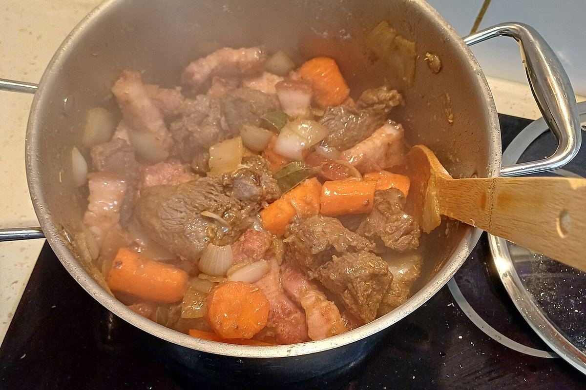 Beef and root vegetables crowded together in one deep pot on a hob, part-way through cooking.