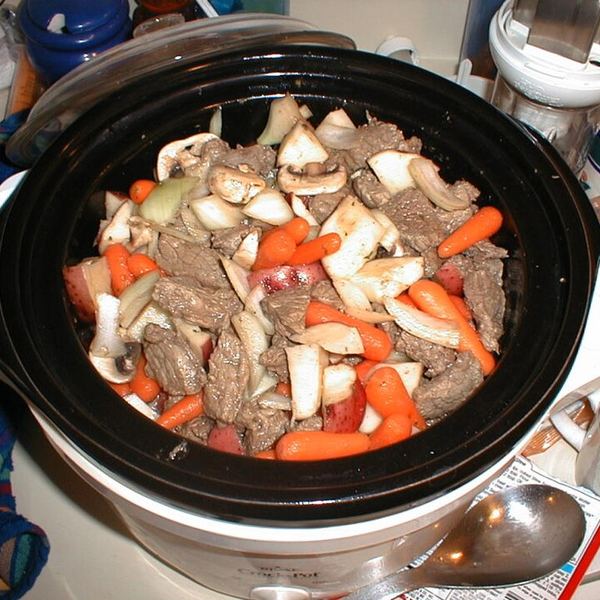 A slow cooker filled with cubes of beef, carrots, onion and mushrooms, before cooking.