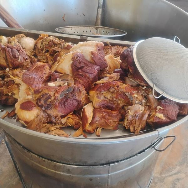 A very large shallow metal cazo full of pieces of pork cooking in fat, with a skimmer resting on the rim.