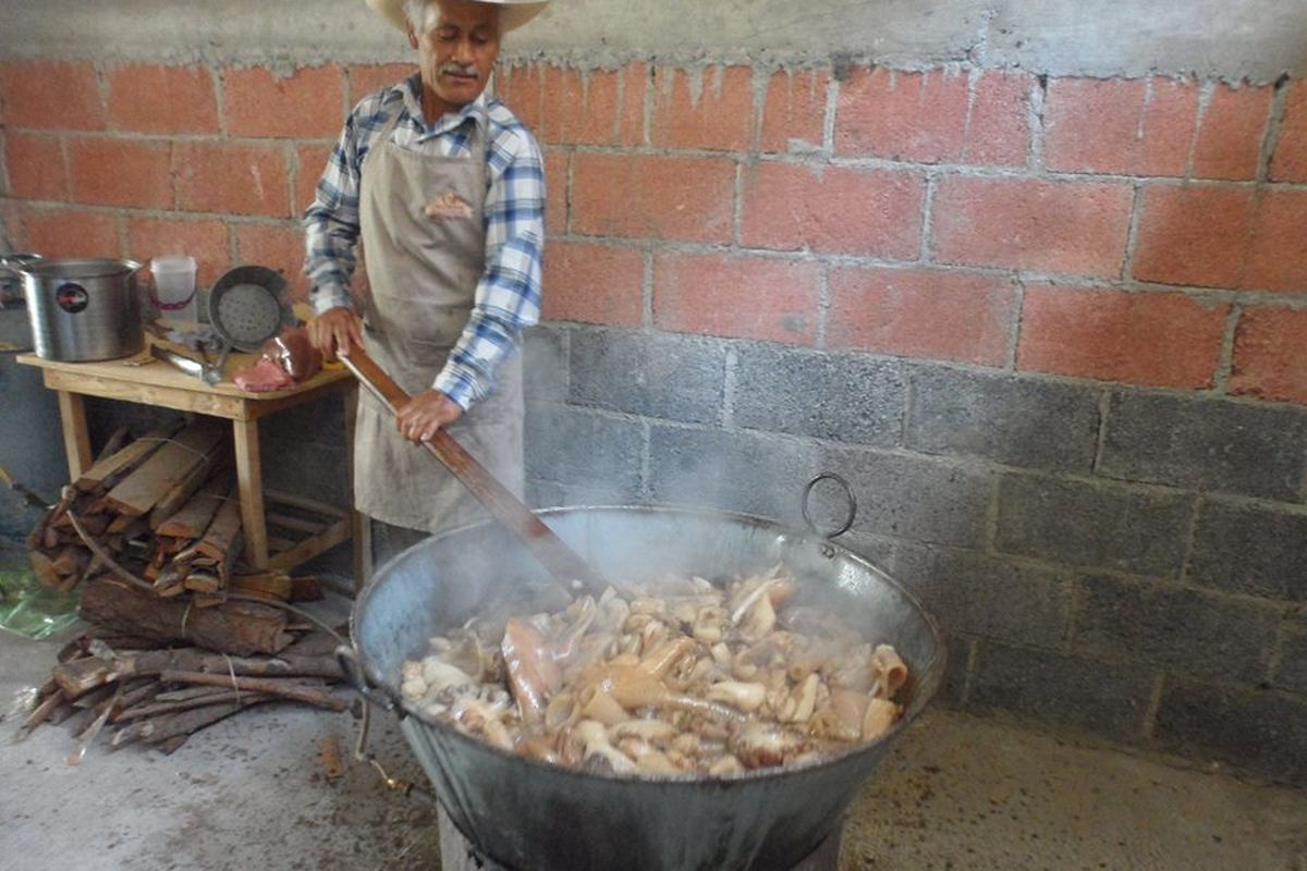 A cook in an apron and straw hat working a very large shallow metal cazo of pork over a wood fire outdoors, stirring with a long wooden paddle.