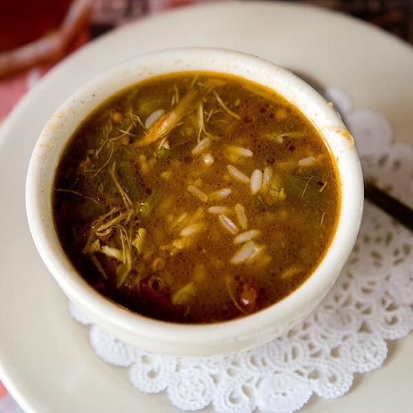 A small white bowl of thin, brothy gumbo with rice, on a saucer.