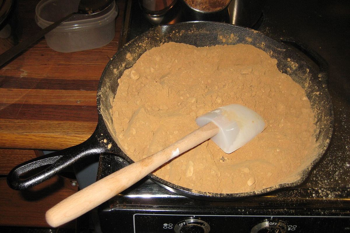 A cast-iron skillet holding a flat bed of toasted flour at peanut-butter colour, with a spatula resting in it.